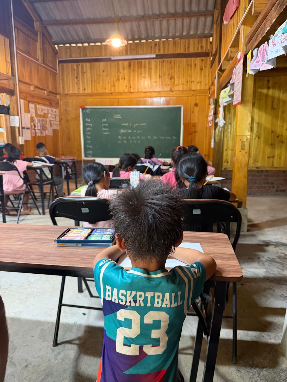 Des enfants au travail à leurs pupitres, vus du fond de la classe en bois.