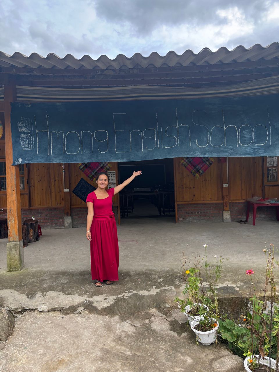Miss Ha en robe rouge, le bras levé vers la bannière en batik indigo où l'on lit « Hmong English School ».