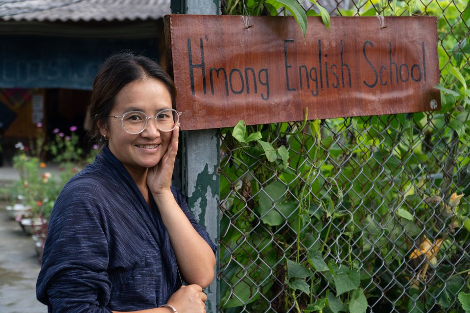 Miss Ha souriante à côté du panneau peint à la main « Hmong English School », à l'entrée du jardin.