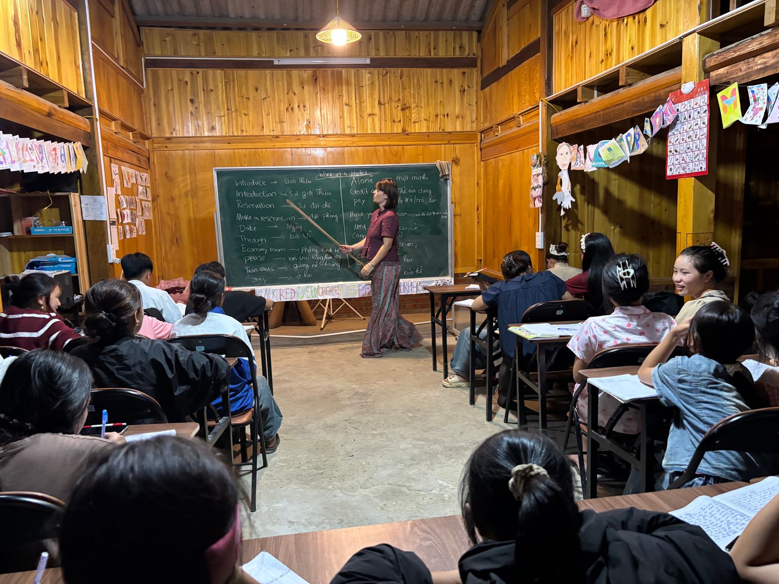 Un bénévole au tableau enseignant le vocabulaire de l'hôtellerie à une classe du soir comble.