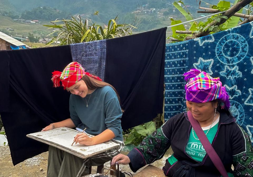 Une invitée dessinant des motifs à la cire chaude aux côtés d'une artisane hmong, des batiks teints à l'indigo suspendus derrière elles.