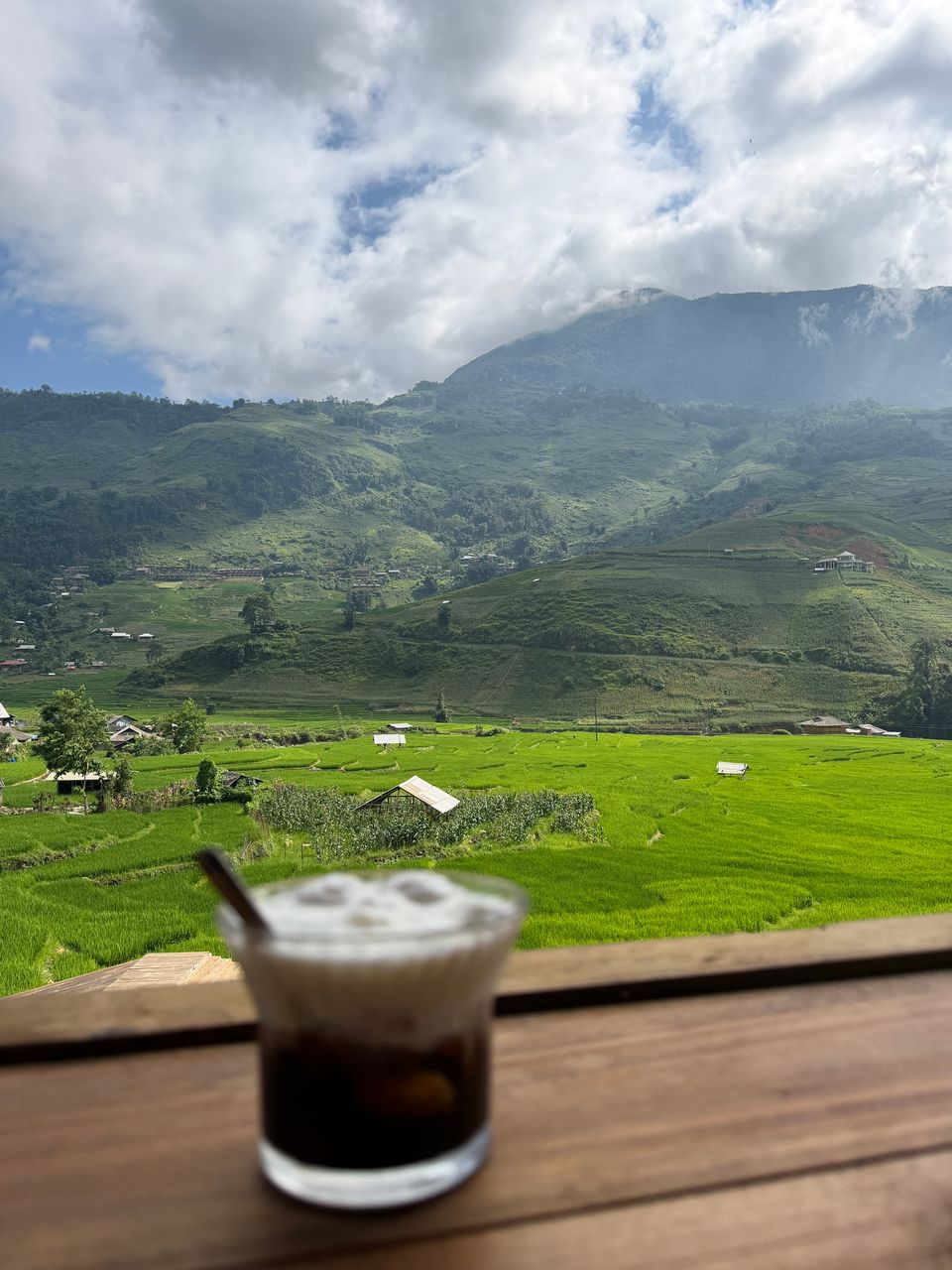 Un café glacé posé sur une rambarde en bois surplombant des rizières d'un vert vif et des montagnes brumeuses.