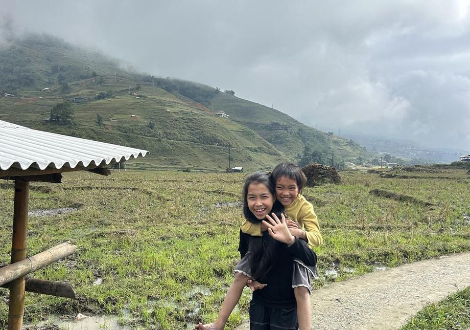 Deux enfants saluant de la main sur un chemin de terre au bord des rizières, l'un sur le dos de l'autre.